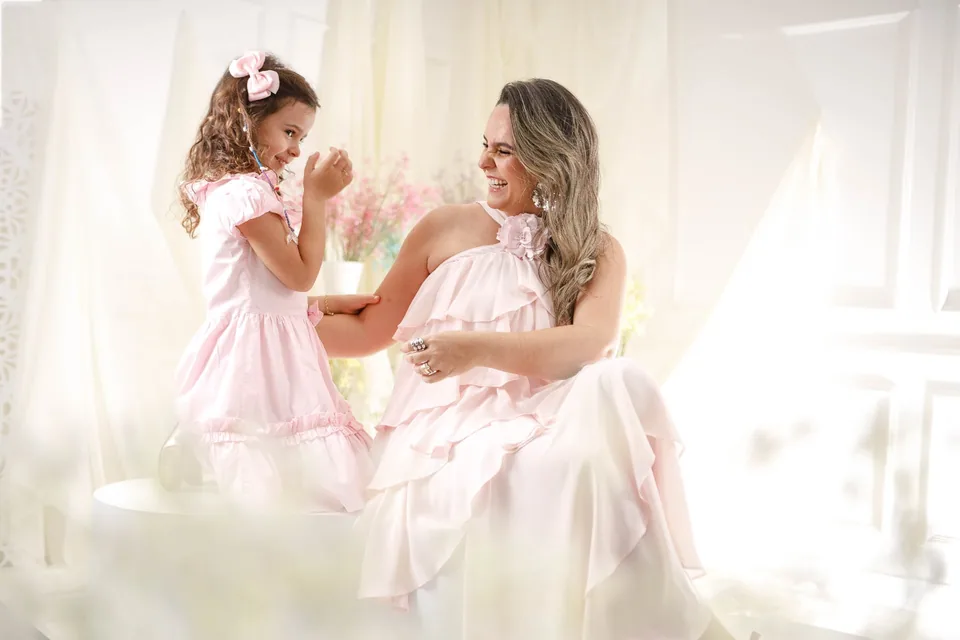Mãe e filha de vestidos rosa sorrindo uma para a outra, em cenário claro e florido