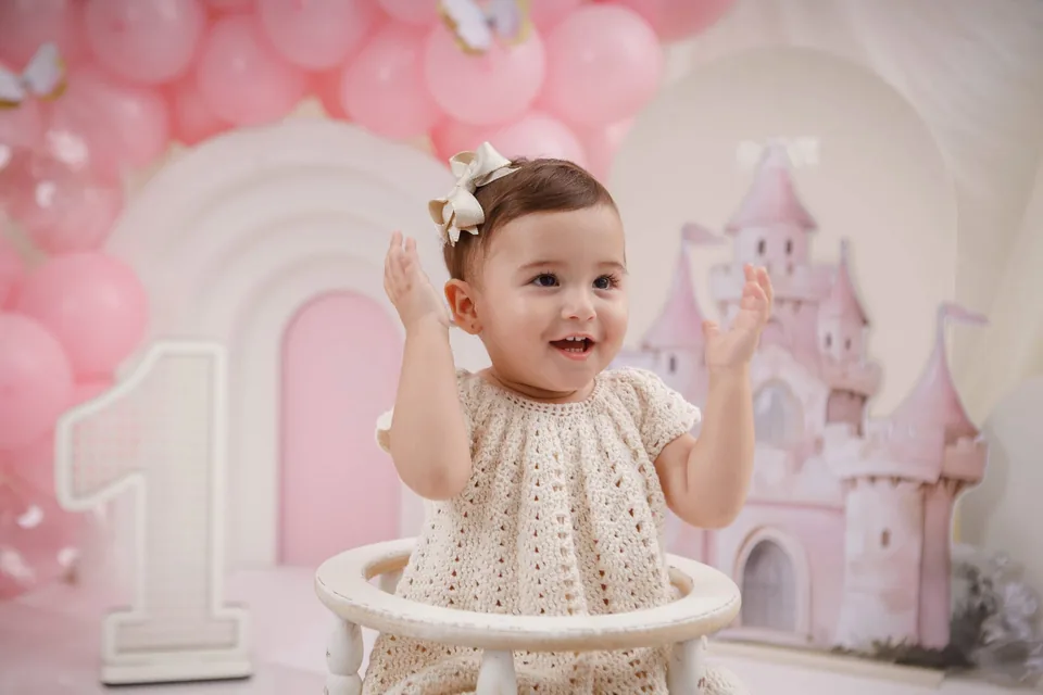 Bebê batendo palminhas e sorrindo, com balões rosa e castelo ao fundo