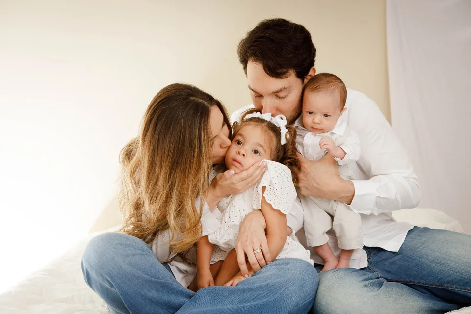 Mãe beijando a filha e pai segurando o bebê, sentados juntos na cama