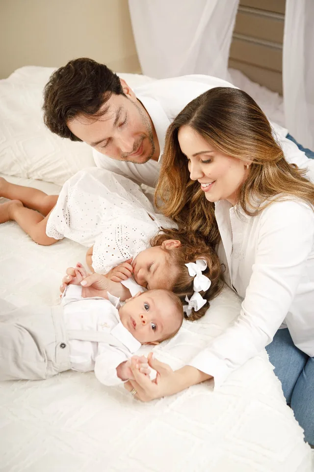 Família de branco deitada na cama, com a filha pequena e o bebê entre os pais