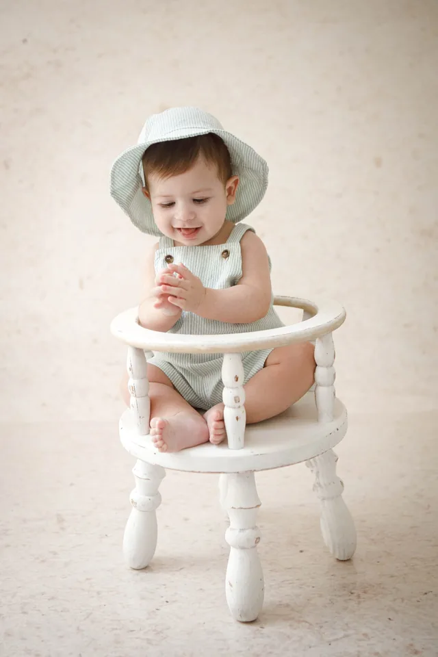 Bebê de chapéu verde brincando com as mãozinhas, sentado em cadeirinha branca