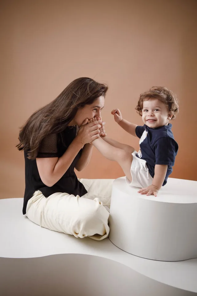Mãe beijando o pezinho do filho, que sorri sentado em um pufe branco