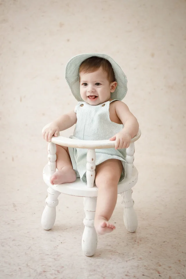 Bebê de chapéu e jardineira verde sorrindo, sentado em cadeirinha branca