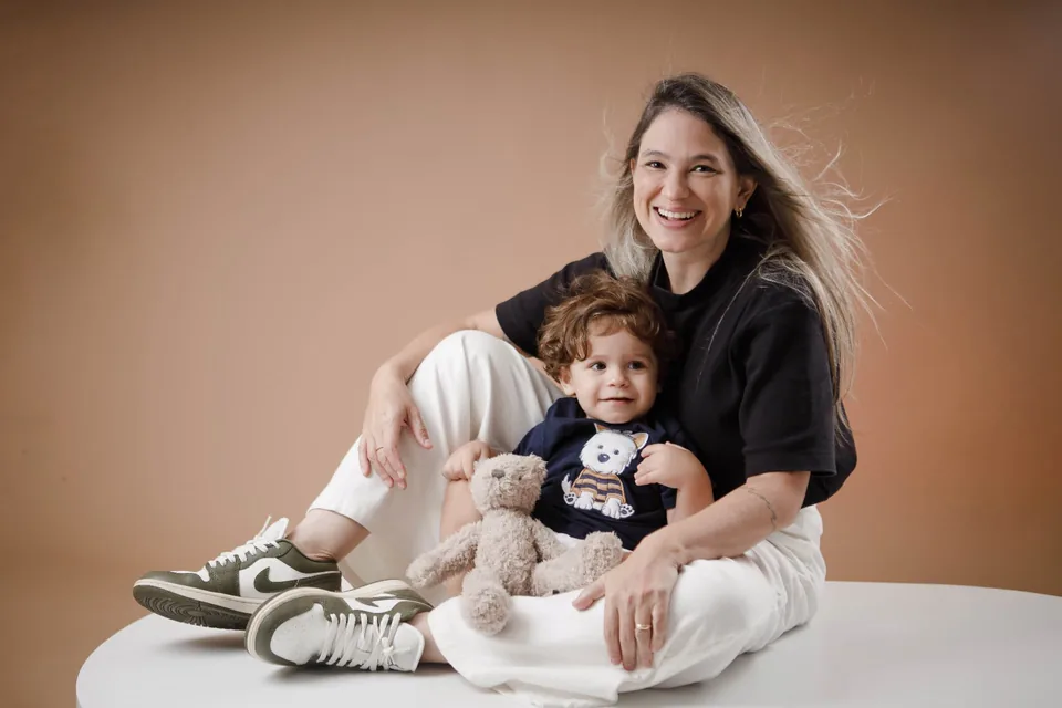 Mãe sentada no estúdio, sorrindo com o filho pequeno e um ursinho de pelúcia no colo
