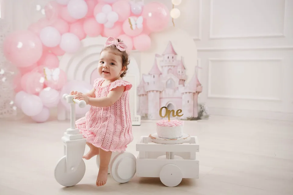 Bebê de vestido rosa em cenário com bolo de primeiro aninho e castelo