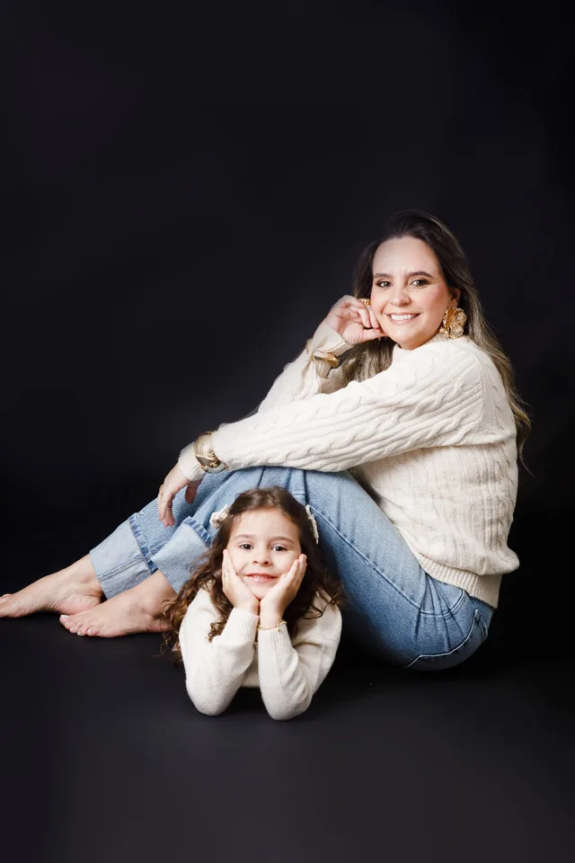 Mãe sorrindo sentada no chão, com a filha apoiando o rosto nas mãos à sua frente