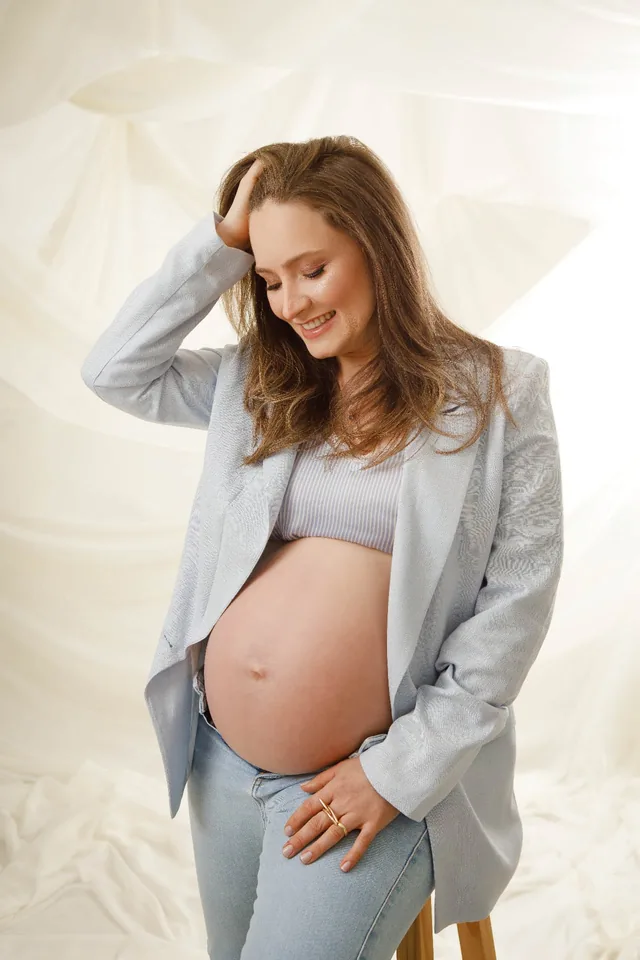 Gestante de blazer azul sorrindo, com a mão nos cabelos e a barriga à mostra, em fundo claro