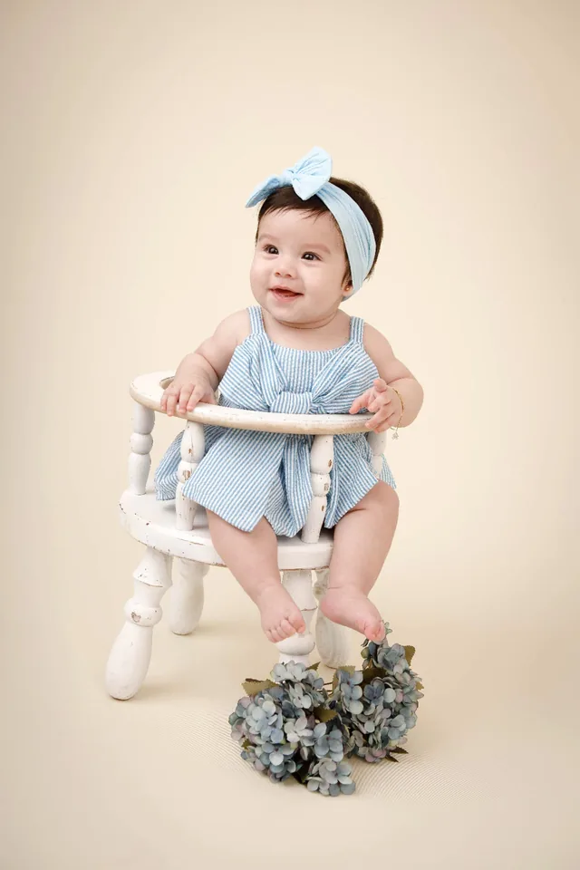 Bebê de vestido listrado azul sentada em cadeirinha branca, com flores aos pés