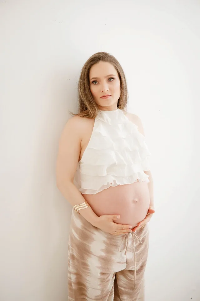 Gestante de blusa branca de babados encostada na parede, com as mãos sobre a barriga