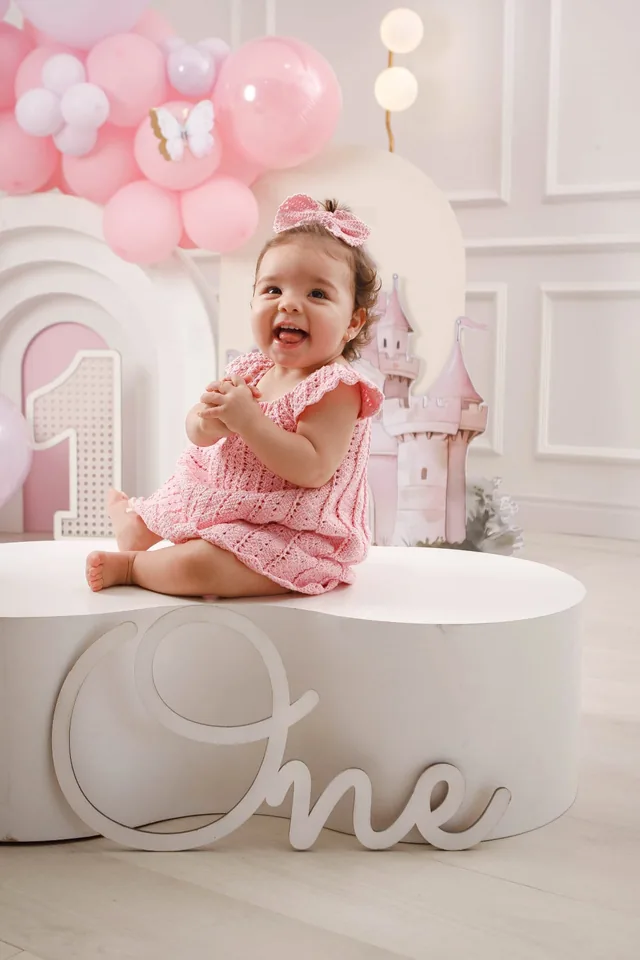 Bebê de vestido rosa sorrindo sobre pufe branco com letreiro “One”