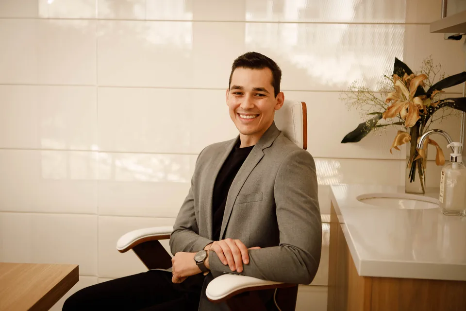 Profissional de blazer cinza sentado em poltrona, sorrindo, em consultório com flores