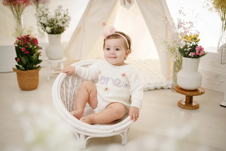 Bebê de blusa “One” sentada em poltroninha de vime, cercada de flores