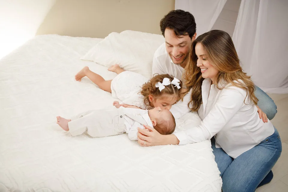 Família de branco deitada na cama, com a filha pequena e o bebê entre os pais