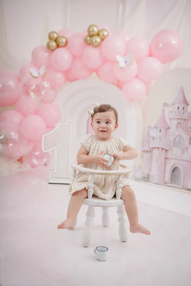 Bebê sentada em cadeirinha branca diante de arco de balões rosa e castelo